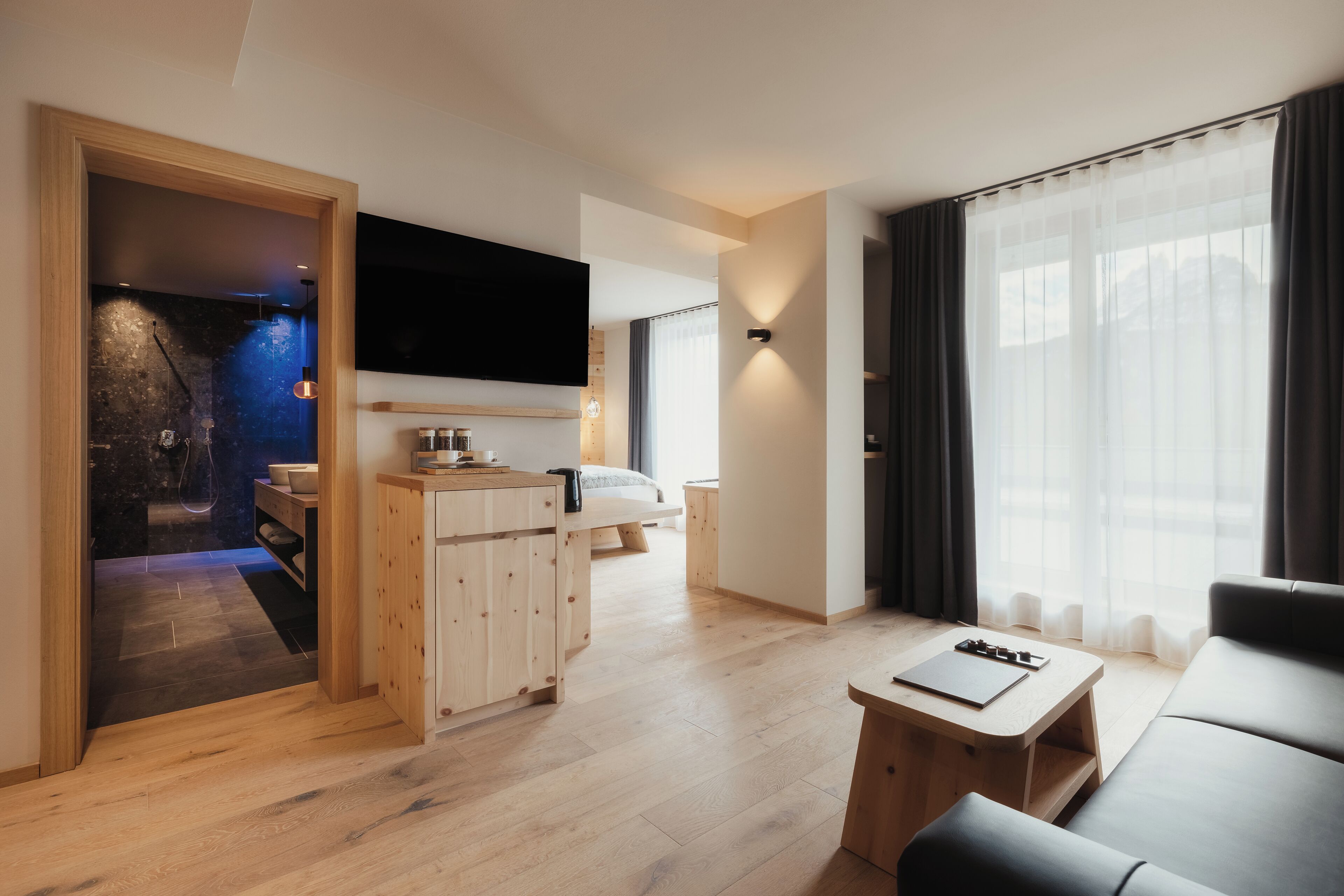 A modern living room with a wooden floor, a black sofa, a small wooden table, and a TV mounted on the wall.