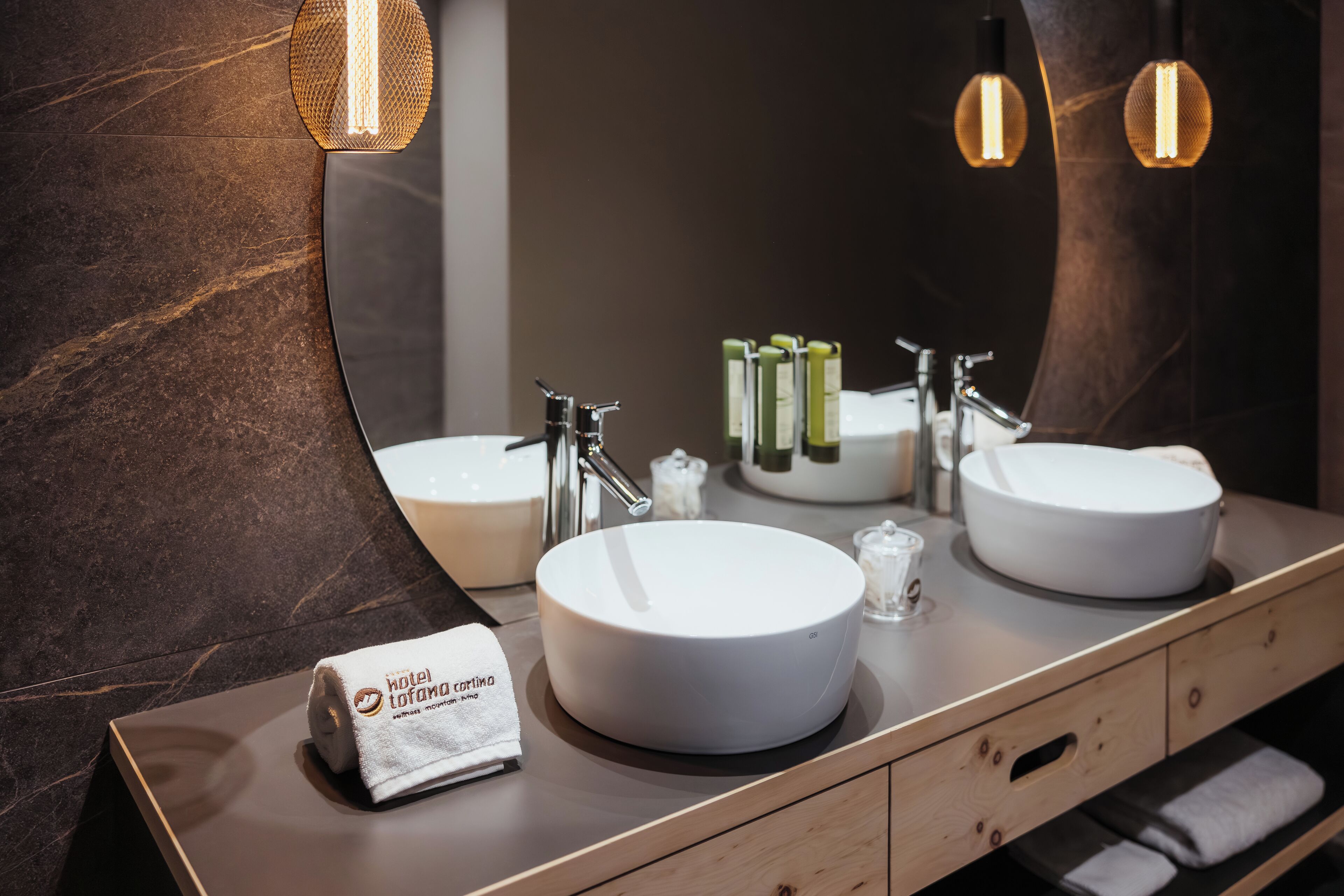 Un baño moderno con dos lavamanos redondos, un espejo grande y un perchero con una toalla de hotel.