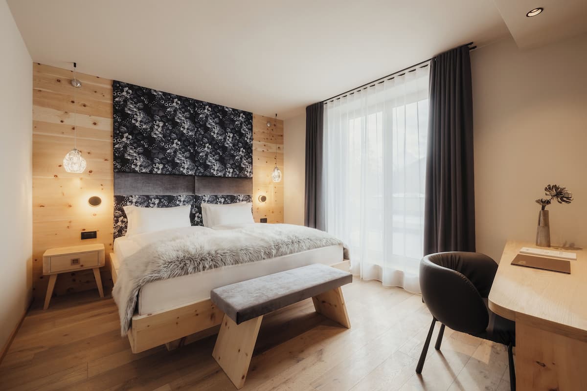 A modern bedroom with a wooden bed, a bench, a desk, and a large window with curtains.