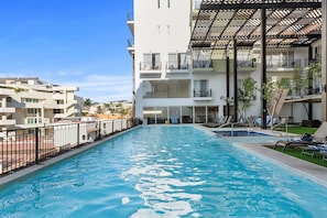 A swimming pool with a clear blue water surface, surrounded by a white building with balconies and a covered outdoor seating area. - 1BR • King Bed • Pool • Jacuzzi • Golden Zone (Bucerías)