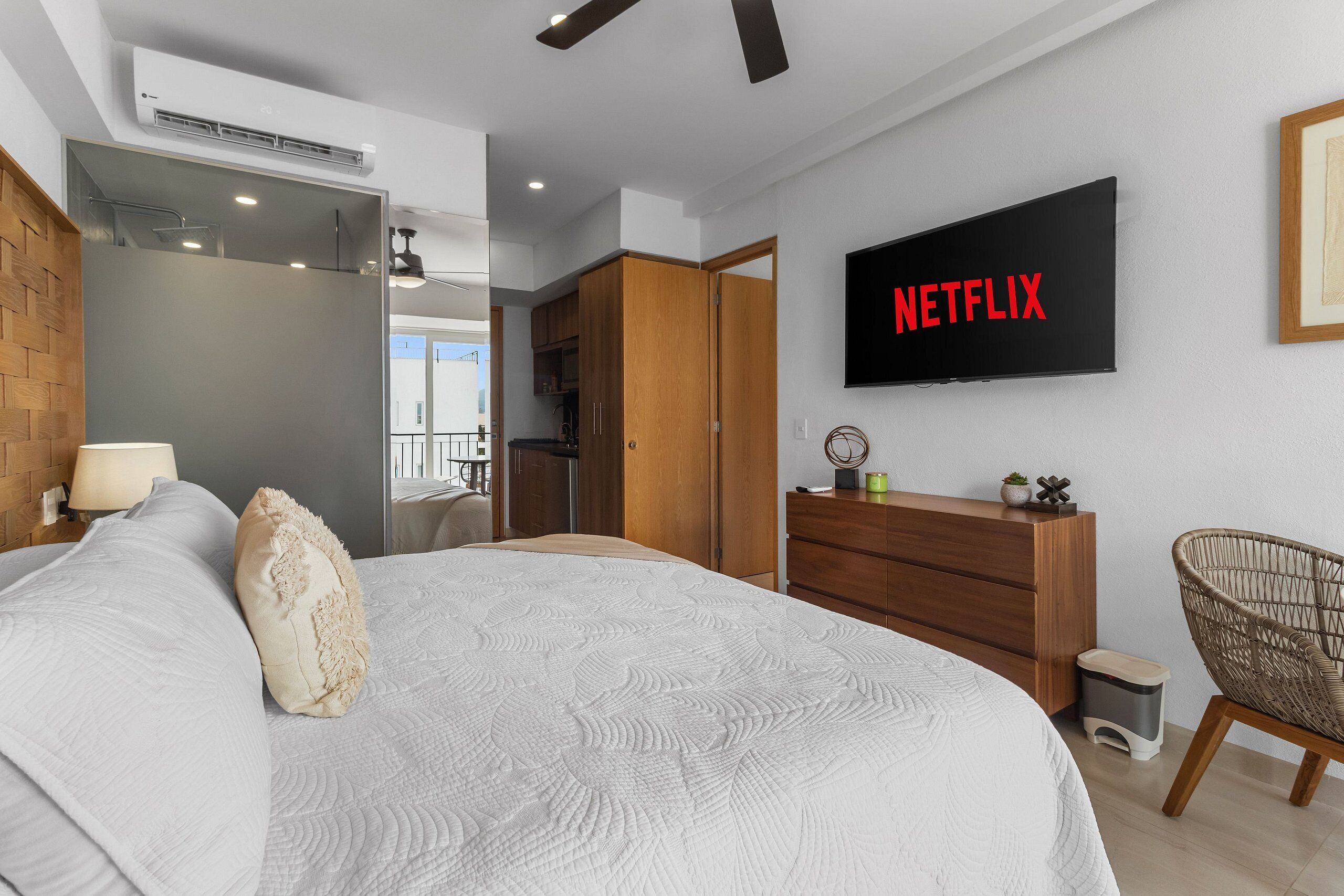 Ett modernt sovrum med en stor säng, en garderob i trä, en tv som visar ”NETFLIX” och en liten papperskorg.