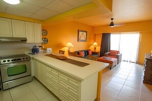 A compact kitchen with a central island, a dining area with a sofa, and a living room with a TV. - Condominio 1HAB/Balcón - Grand Marina Villas (Nuevo Vallarta)