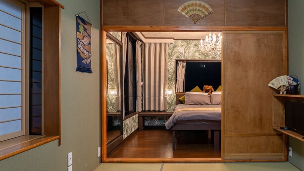 A traditional Japanese-style room with tatami flooring, a sliding door, a bed with pillows, a fan, and a small shelf with decorative items.
