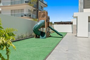 Une aire de jeux avec un toboggan et une structure d’escalade, entourée d’une pelouse artificielle verte et d’un mur blanc.