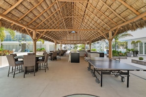 Un espace extérieur aménagé avec des tables et des chaises, une table de ping-pong et une structure dotée d’un toit de chaume.