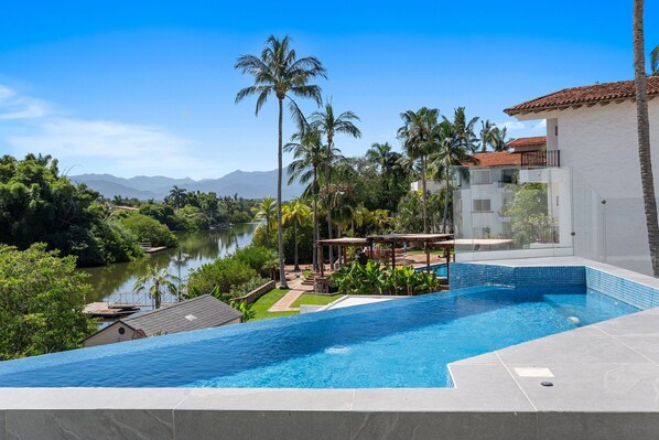 A swimming pool with a view of a river and mountains, surrounded by palm trees and a building with a tiled roof.