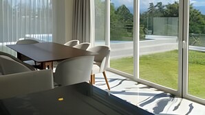 A modern dining area with a large table and chairs, large windows with curtains, and a view of a road and greenery. - 2BR Exclusive Private Pool • Canal view y Beach (Las Jarretaderas)