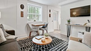 Un salon moderne comprenant un canapé, une table basse, un coin repas avec une table et des chaises, un téléviseur fixé au mur et une fenêtre avec des stores.