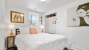 Une chambre avec un grand lit, deux tables de chevet avec des lampes, une fenêtre avec des stores, un tableau encadré au mur et une porte donnant sur une autre pièce.