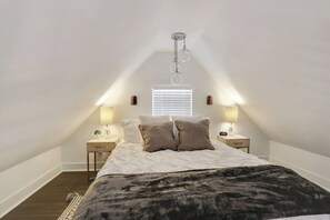 A bedroom with a vaulted ceiling, a bed with a dark blanket, two bedside tables with lamps, and a window with blinds.