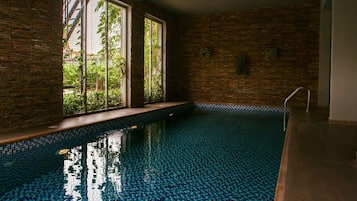 Una piscina interior con suelo de baldosas, amplias ventanas con vistas a zonas verdes y una pared de ladrillo.