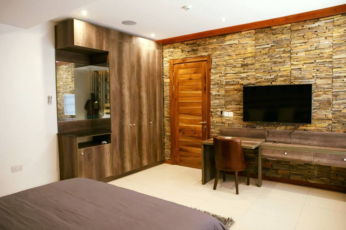 A bedroom with a bed, a wooden wardrobe, a TV mounted on a stone wall, and a small desk with a chair.