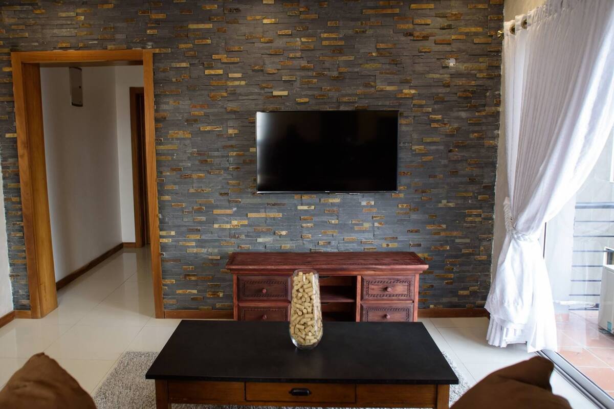 A living room with a flat-screen TV mounted on a brick-patterned wall, a wooden cabinet, and a table with a decorative vase.