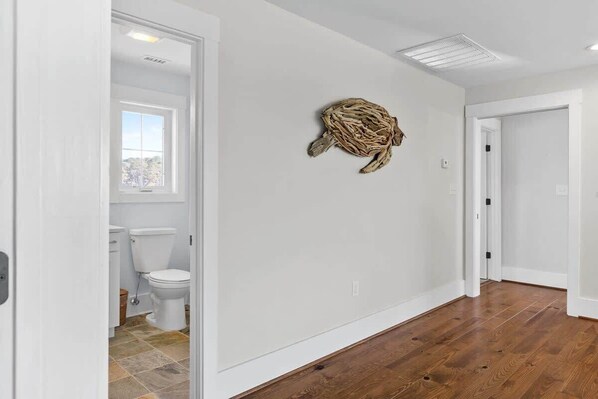 Un pasillo con suelo de madera, una pared blanca con un adorno y vistas a un baño con inodoro y ventana.