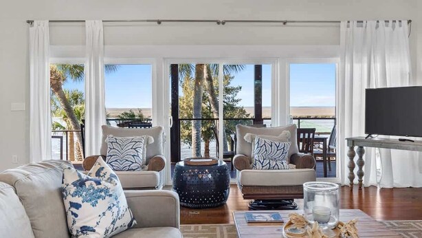 A living room with a sofa, two armchairs, a coffee table, and a TV. There are large windows with a view of palm trees and a beach.