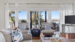 Una sala de estar con sofá, dos sillones, una mesa de centro y televisor. Se observan amplias ventanas con vista a palmeras y una playa.