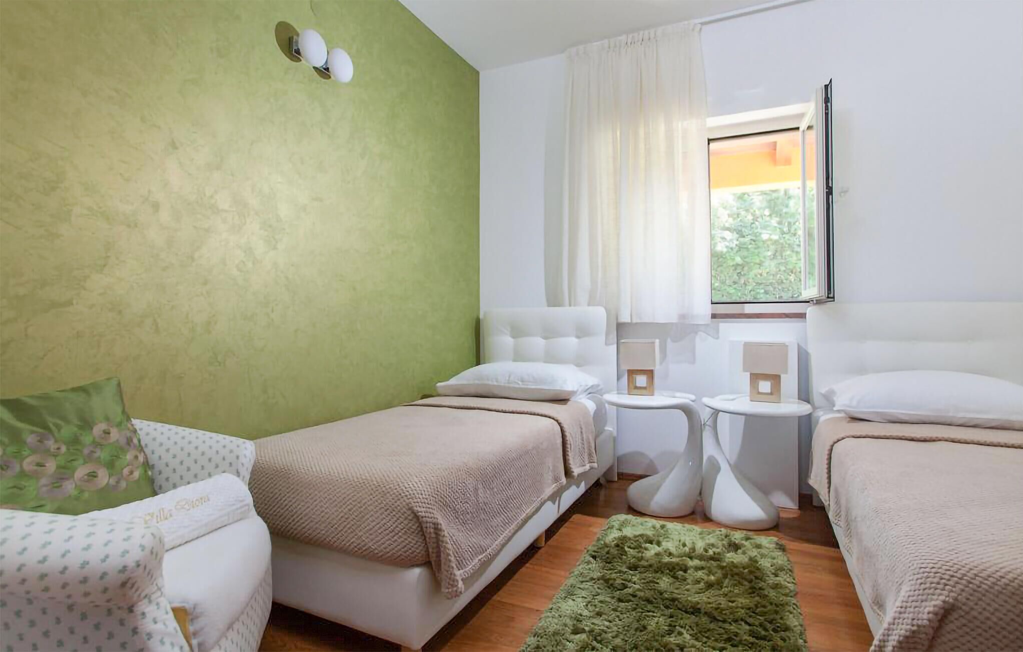 A room with two beds, a green accent wall, a window with curtains, and a small table with a lamp.