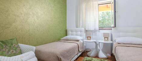 A room with two beds, a green accent wall, a window with curtains, and a small table with a lamp.