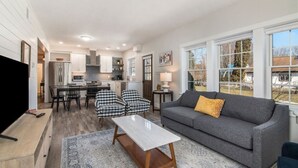 Ein modernes Wohnzimmer mit einem grauen Sofa, einem Couchtisch und karierten Stühlen. Im Hintergrund ist eine Küche mit weißen Schränken und ein Essbereich mit Tisch und Stühlen zu sehen.