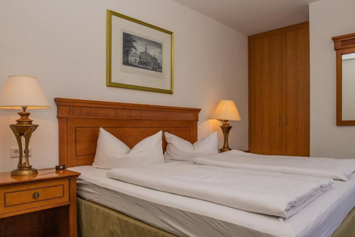 A hotel room with a wooden headboard, two beds, a nightstand with a lamp, a framed picture on the wall, and a wardrobe.