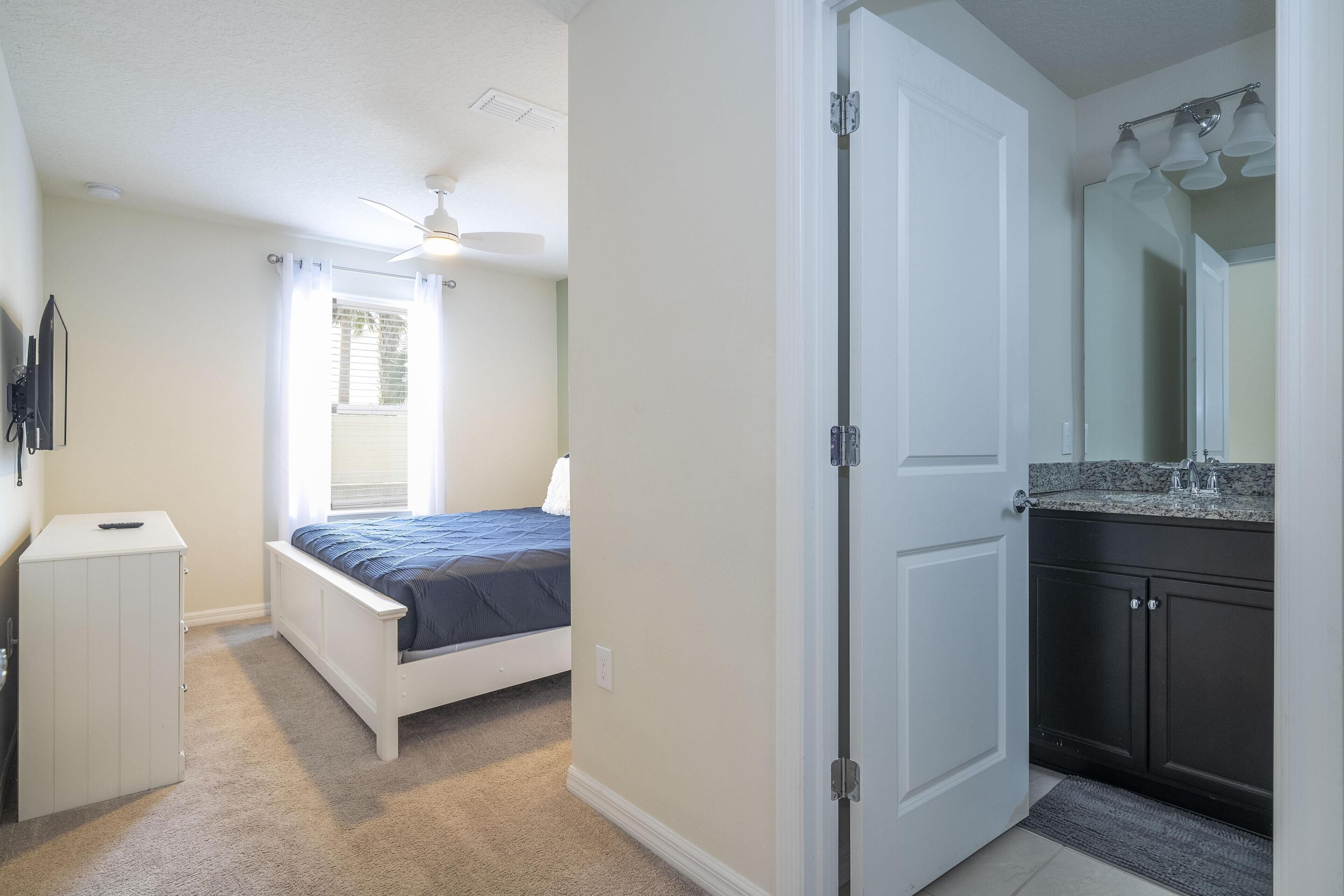 A bedroom with a bed, a ceiling fan, a window with curtains, a white wardrobe, and a bathroom with a sink and mirror.