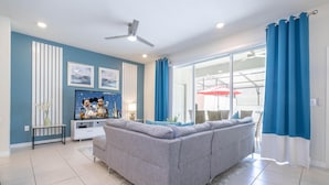 Een woonkamer met een grijze bank, een plafondventilator en een groot raam met blauwe gordijnen.
