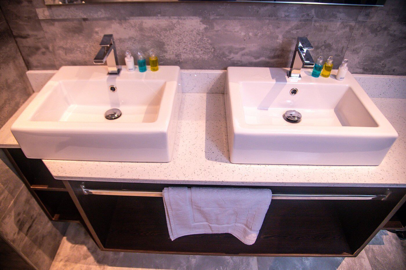 A bathroom with two white sinks, each with a faucet and soap dispensers. There is a towel hanging on a rack below the sinks.
