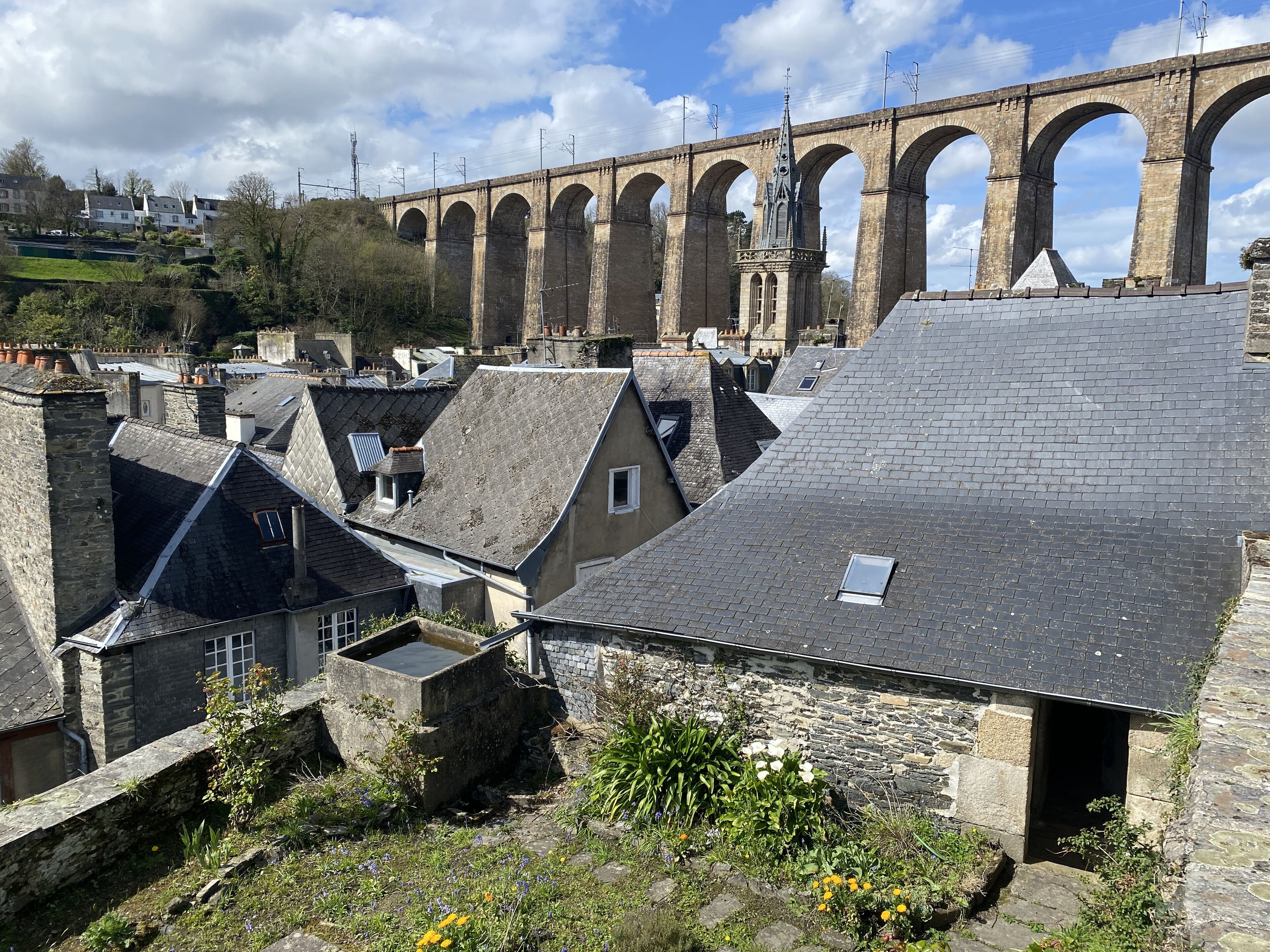 A historic stone bridge with multiple arches, a stone building with a sloped roof, and a small garden area with flowers.