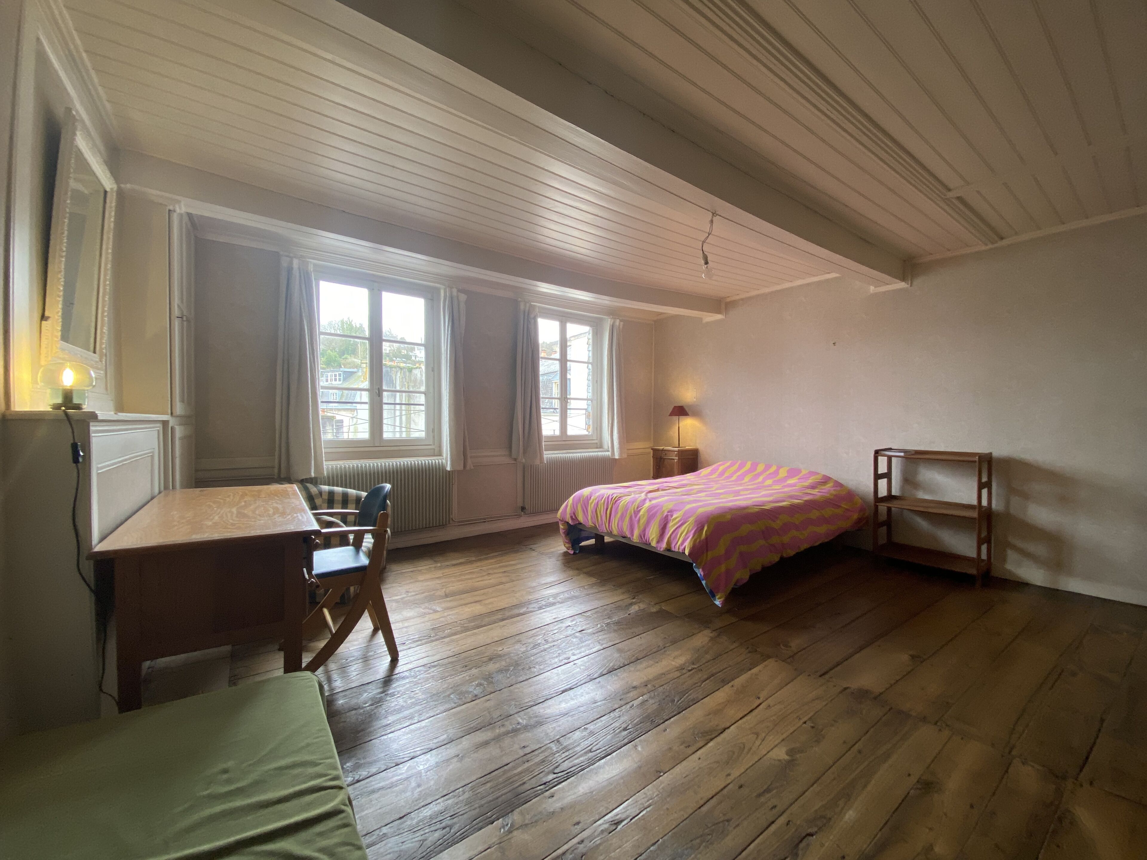A room with a wooden floor, a bed with a striped bedspread, a small table with chairs, and two windows with curtains.