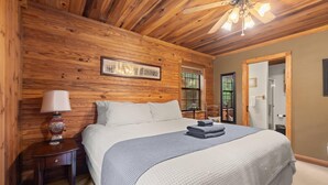 Une chambre avec un plafond en bois, un lit recouvert d’une couverture bleue et blanche, une table de chevet avec une lampe et un tableau encadré accroché au mur.