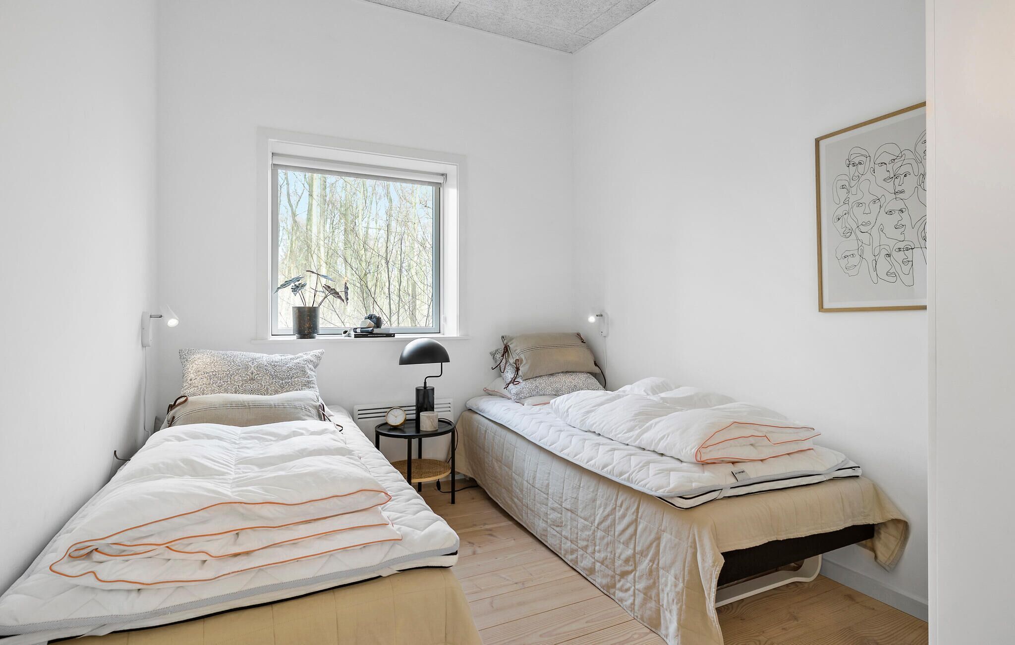 Una camera da letto piccola e luminosa con due letti singoli, una finestra vista alberi, un comodino con una lampada e un quadro astratto incorniciato appeso al muro.