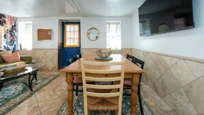 Ein Essbereich mit einem Holztisch, Stühlen und einem Teppich. Eine blaue Tür, ein Spiegel an der Wand und ein Fenster mit Vorhängen sind zu sehen.