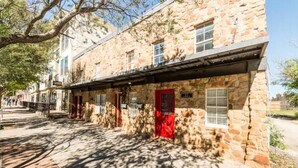 A stone building with a red door, a covered walkway, and a tree casting shadows on the ground. - Industrial St. Loft on the square (Denton)