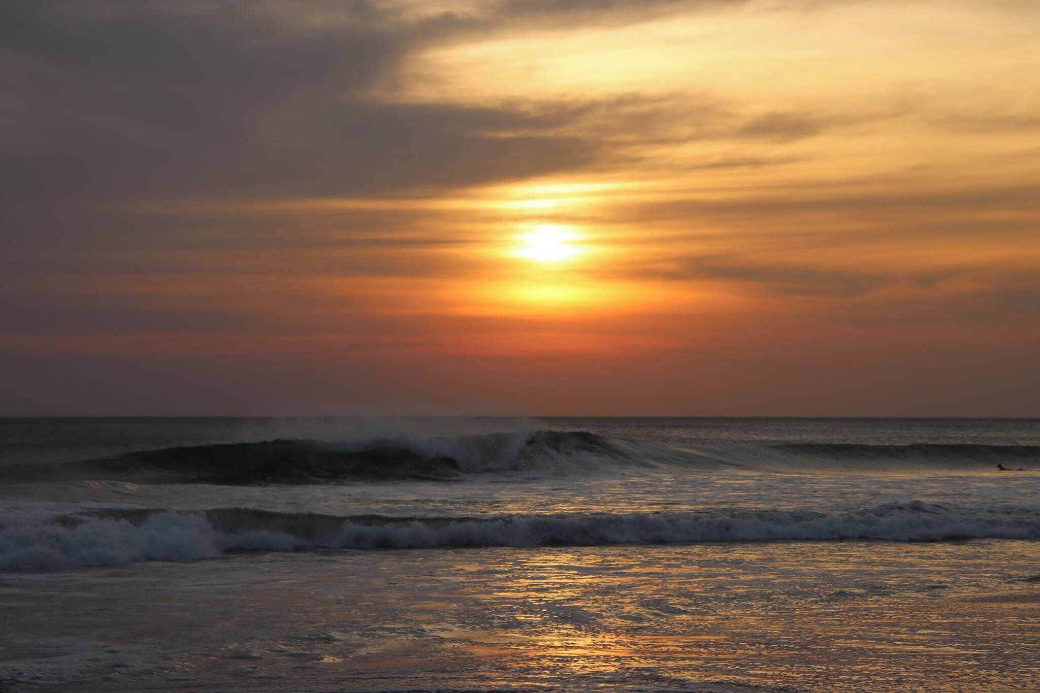 A sunset over the ocean with waves and a person surfing.