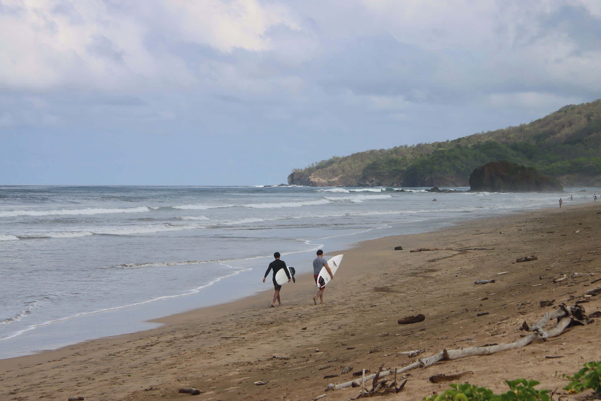 A beach with two people carrying surfboards, a sandy shore, and a forested hill in the background.