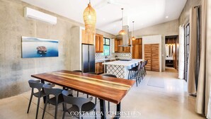 Una cocina moderna con armarios de madera, una isla central, iluminación colgante y una zona de comedor con mesa y sillas de madera.
