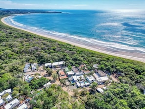 An aerial view of a coastal area with a beach, dense greenery, and a cluster of buildings with flat roofs.