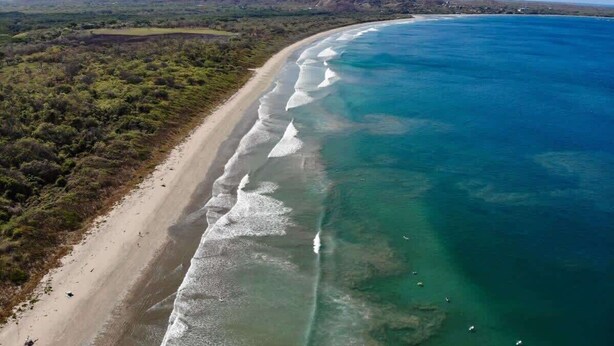 An aerial view of a beach with clear waters, a sandy shore, and a hilly landscape in the background.