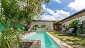 A swimming pool surrounded by tropical plants and a modern building with a covered patio area.