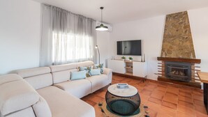 A modern living room with a beige sofa, a round coffee table, a stone fireplace, and a flat-screen TV mounted on the wall. - Casa Mati Cómpeta by Ruralidays (Málaga)