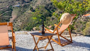 A wooden table with a bottle and glass, a straw hat, and a chair on a gravel surface with a view of a hilly landscape. - Casa Mati Cómpeta by Ruralidays (Málaga)