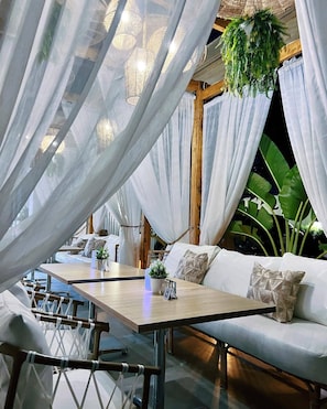 Un espace de restauration intérieur avec une table en bois, des sièges rembourrés de couleur blanche et des rideaux légers.