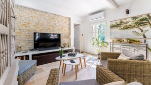 A modern living room with a stone wall, a flat-screen TV, wicker furniture, and a large window. - Provencal bastide with heated pool near Alpilles and Luberon (Vernègues)