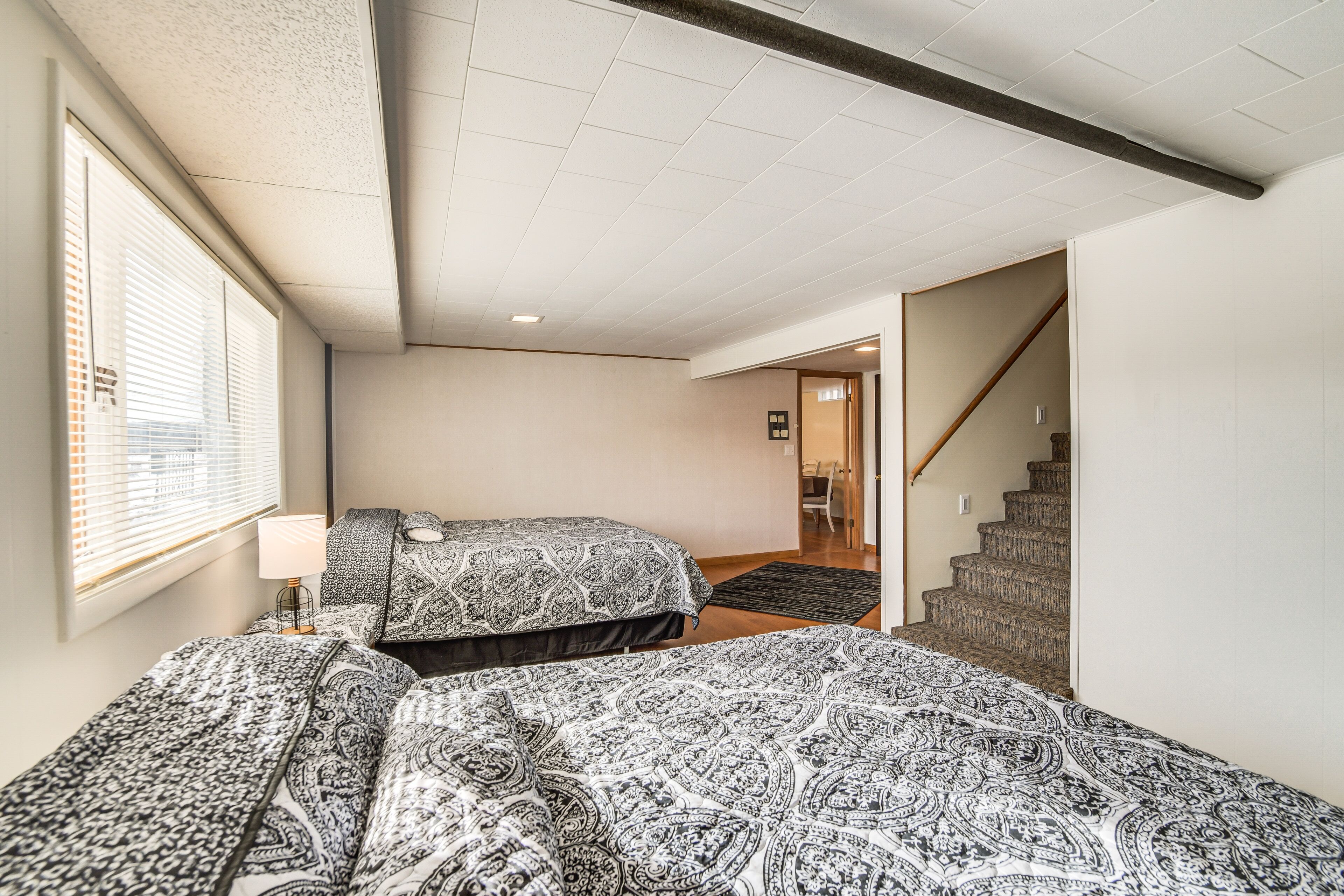 A bedroom with two beds, a window with blinds, a staircase, and a door leading to another room.