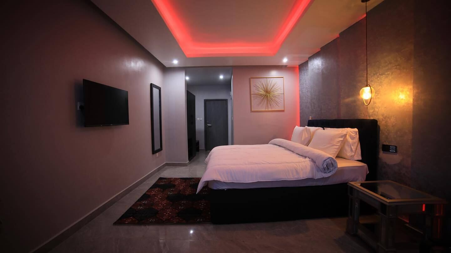 A modern bedroom with a bed, a bedside table, a TV mounted on the wall, and a door leading to another room.