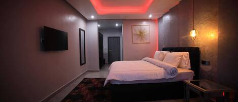 A modern bedroom with a bed, a bedside table, a TV mounted on the wall, and a door leading to another room.