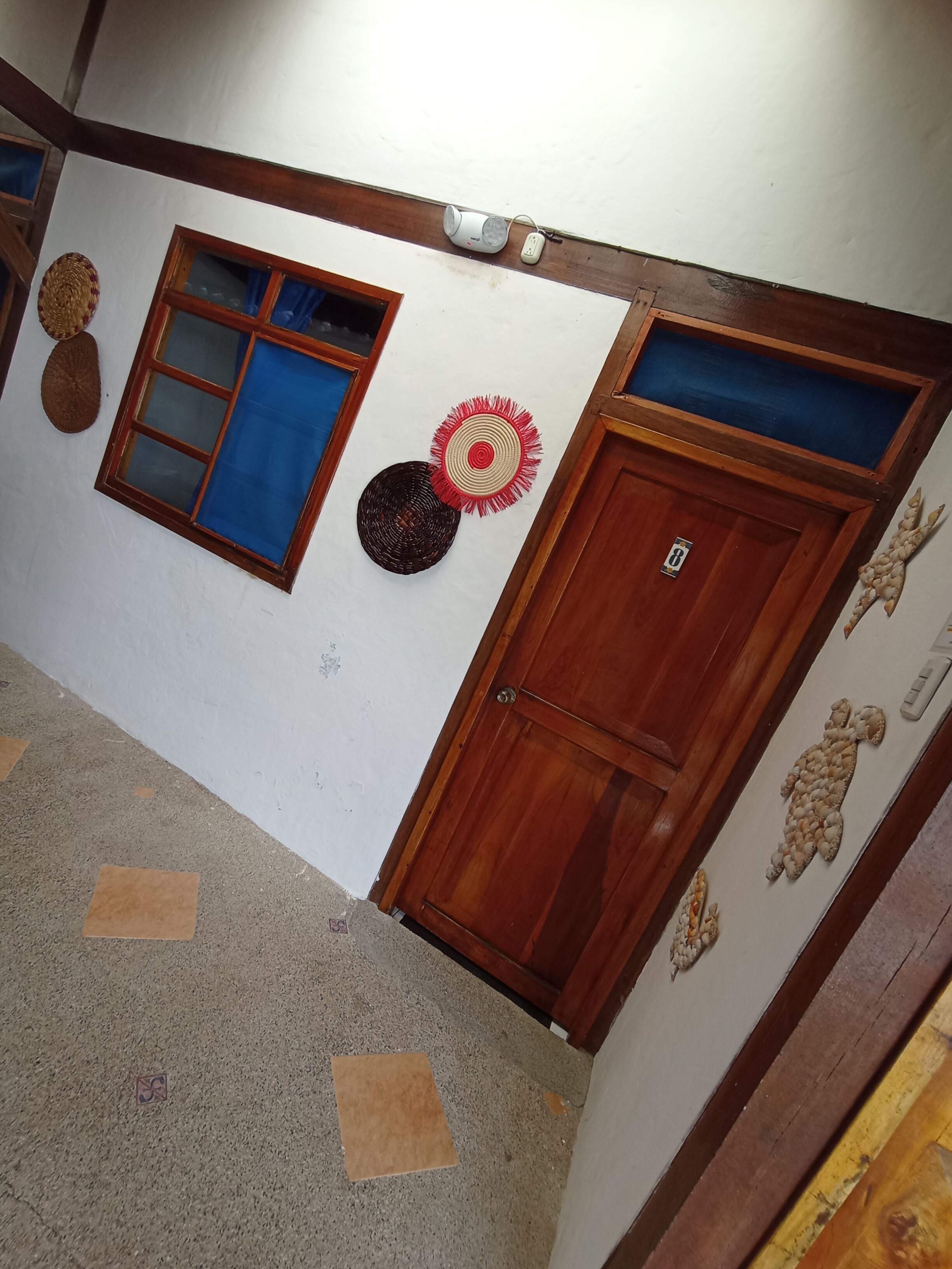 Un pasillo con una puerta de madera, una ventana con cortinas azules y adornos en la pared, incluyendo una cesta tejida y un objeto circular.