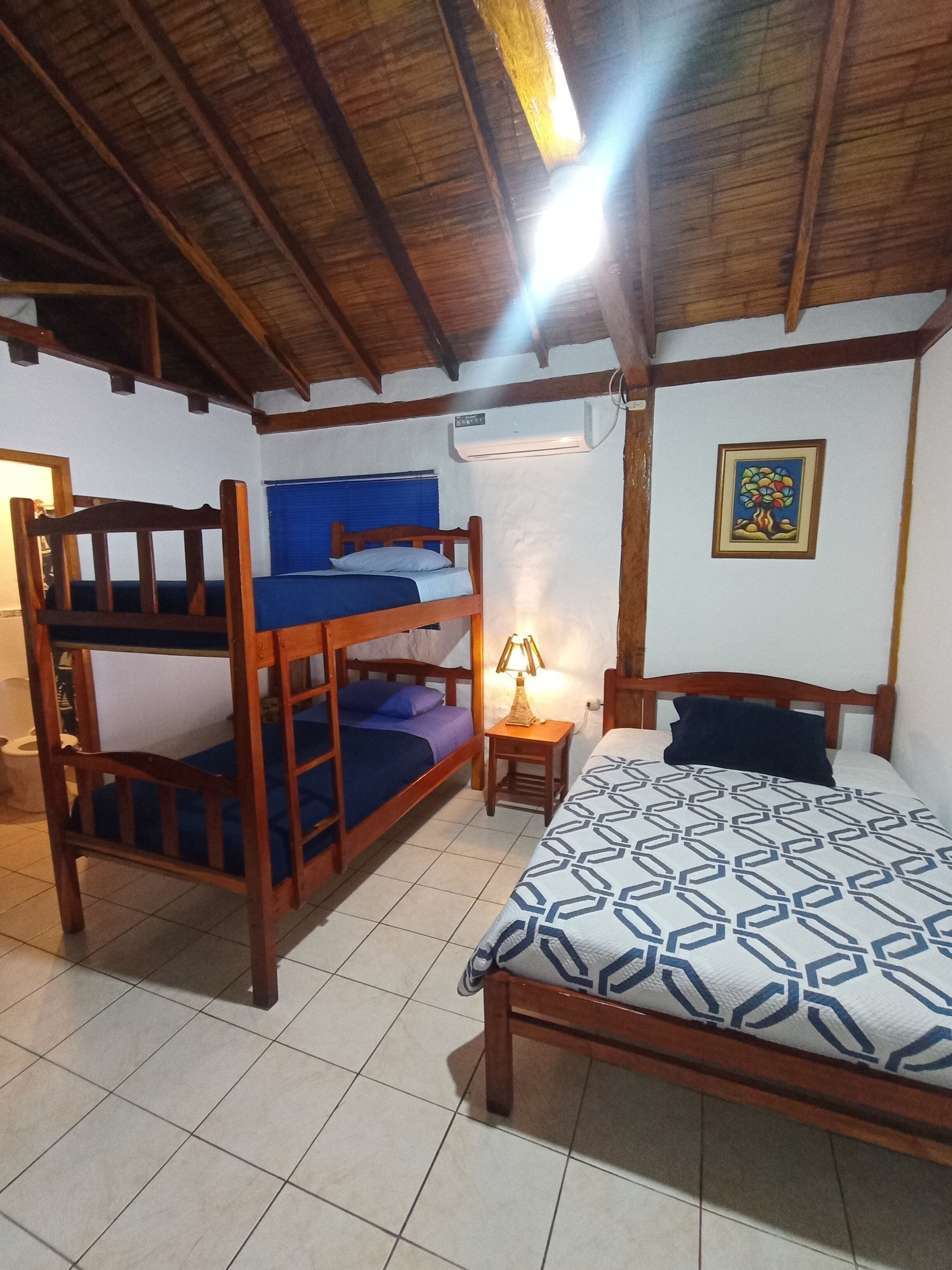 Una habitación con una cama litera, una cama individual, un equipo de aire acondicionado, una mesita con una lámpara y un cuadro en la pared.