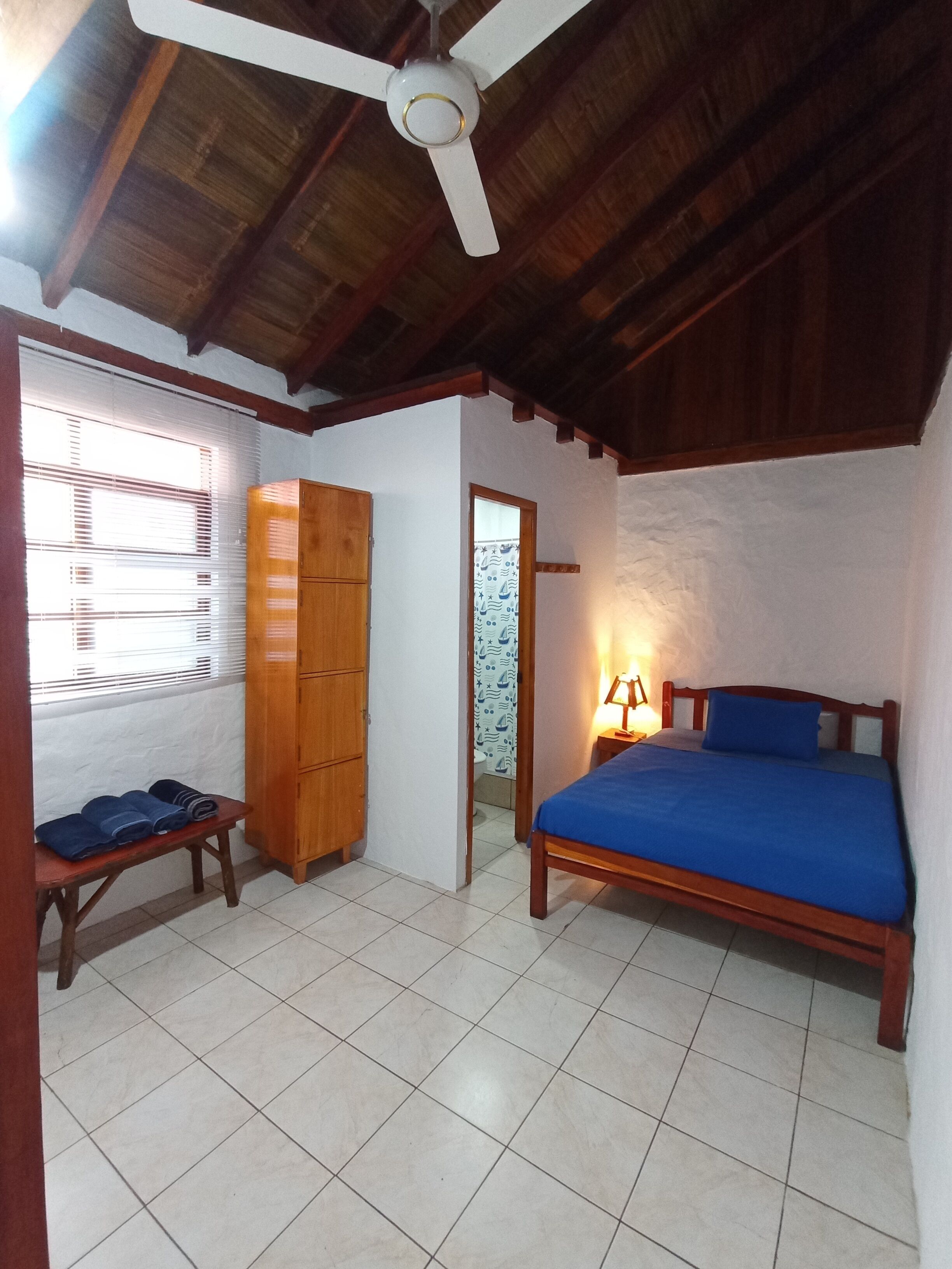 Un dormitorio con ventilador de techo de madera, una cama con colchón azul, un armario de madera y un banco con toallas.