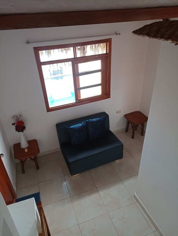A living room with a dark blue sofa, a small wooden side table with a vase of flowers, a window with wooden frames, and a tiled floor. - Hotel SoleMare (Manglaralto)